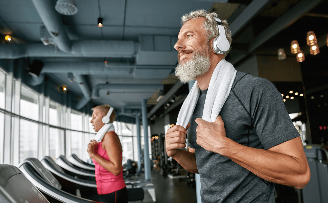 Man with grey hair and beard with headphones on walking on a treadmill.